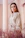 A smiling woman wearing a beige rosé caftan with delicate embroidery, standing in a softly lit room.