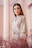 A smiling woman wearing a beige rosé caftan with delicate embroidery, standing in a softly lit room.