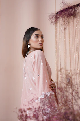 A graceful woman draped in a soft pastel pink silk saree standing against a minimalist white backdrop.