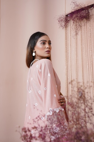 A woman stands gracefully at an angle, wearing a delicate pink outfit adorned with white floral embroidery and lacework. Her long hair falls softly over her shoulders, and she wears elegant white earrings. The background features soft pink hues and ethereal dried flowers, creating a serene and sophisticated atmosphere.