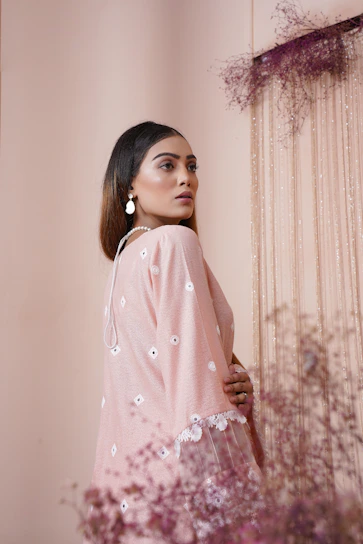 Elegant woman wearing a soft blush pink matching top and skirt set, standing by a sunlit window.