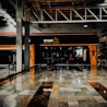 A fitness center entrance with the branding 'smart fit' illuminated above the doors. The interior has a modern design, featuring dark glass and bright orange accents. A reflective marble floor extends in front, capturing reflections from the surroundings. There are two individuals standing and walking towards the left.