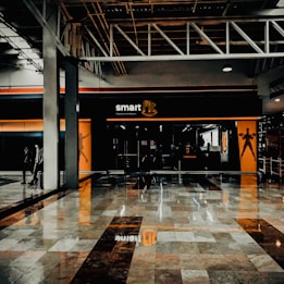 A fitness center entrance with the branding 'smart fit' illuminated above the doors. The interior has a modern design, featuring dark glass and bright orange accents. A reflective marble floor extends in front, capturing reflections from the surroundings. There are two individuals standing and walking towards the left.