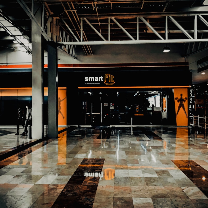A fitness center entrance with the branding 'smart fit' illuminated above the doors. The interior has a modern design, featuring dark glass and bright orange accents. A reflective marble floor extends in front, capturing reflections from the surroundings. There are two individuals standing and walking towards the left.