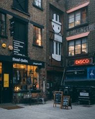 A narrow alleyway features a cozy cafe with signage reading 'Bread & Butter' and 'playground72'. Outdoor seating includes a small table and chairs. Above the cafe, black signs display options such as coffee, workspace, and events. Opposite the cafe, there's a large mural of a stylized character in black and white on a brick wall. A neon sign that reads 'DISCO' is affixed to a wall above a commercial waste bin, further contributing to the eclectic urban ambiance. The walls are adorned with graffiti, and the ground is paved with light-colored bricks.