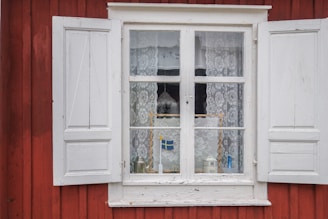 A window with white wooden shutters on a red wooden building features lace curtains and various objects displayed on the windowsill, including a small Swedish flag and decorative items.