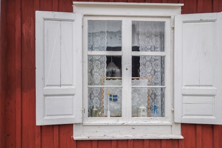 A window with white wooden shutters on a red wooden building features lace curtains and various objects displayed on the windowsill, including a small Swedish flag and decorative items.