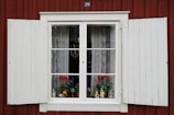 A vintage-style window with white wooden shutters on a red wooden wall. The window displays lace curtains, potted red flowers, and small decorative items including figurines and a candle holder.