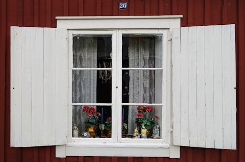 A vintage-style window with white wooden shutters on a red wooden wall. The window displays lace curtains, potted red flowers, and small decorative items including figurines and a candle holder.