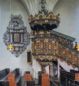 An ornate church interior with a richly decorated pulpit featuring intricate carvings and colorful details. The pulpit is adorned with gold accents and various religious motifs. Wooden pews are arranged neatly, and a decorative plaque with an elaborate black and white design is mounted on the wall. The lighting is soft, and the overall atmosphere is serene and historical.