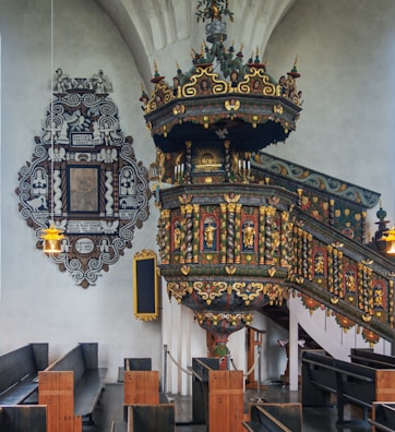An ornate church interior with a richly decorated pulpit featuring intricate carvings and colorful details. The pulpit is adorned with gold accents and various religious motifs. Wooden pews are arranged neatly, and a decorative plaque with an elaborate black and white design is mounted on the wall. The lighting is soft, and the overall atmosphere is serene and historical.