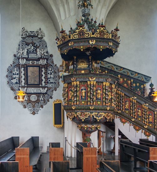 An ornate church interior with a richly decorated pulpit featuring intricate carvings and colorful details. The pulpit is adorned with gold accents and various religious motifs. Wooden pews are arranged neatly, and a decorative plaque with an elaborate black and white design is mounted on the wall. The lighting is soft, and the overall atmosphere is serene and historical.