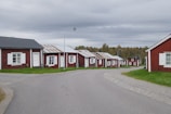A row of small red wooden houses with white trim lines a gently curving paved road. The houses have metal roofs, some appearing weathered. Each house has a small front yard with green grass. Overhead, a cloudy gray sky creates a calm atmosphere, and in the background, there is a treeline with autumn-colored foliage.