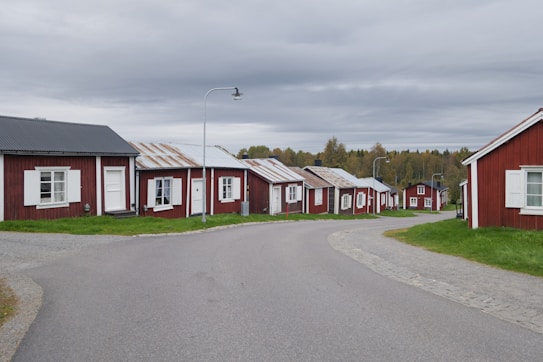 A row of small red wooden houses with white trim lines a gently curving paved road. The houses have metal roofs, some appearing weathered. Each house has a small front yard with green grass. Overhead, a cloudy gray sky creates a calm atmosphere, and in the background, there is a treeline with autumn-colored foliage.