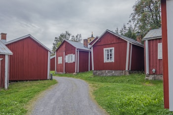 Several small, red wooden cottages with white trim situated along a gravel pathway, surrounded by green grass and trees on a cloudy day.