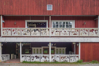 A two-story wooden structure with red paneling and intricate white decorative railings featuring various cutout patterns. The upper level includes a large window and recessed interior space with hanging string lights. There are multiple windows on both levels and lantern-style outdoor lights mounted on the walls.