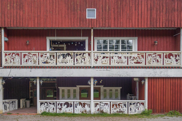 A two-story wooden structure with red paneling and intricate white decorative railings featuring various cutout patterns. The upper level includes a large window and recessed interior space with hanging string lights. There are multiple windows on both levels and lantern-style outdoor lights mounted on the walls.