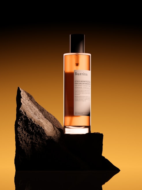 A perfume bottle with a black cap and an amber liquid inside is placed on a rough, dark rock slab. The background has a gradient from dark at the top to a lighter brownish-orange hue at the bottom, creating a dramatic contrast.