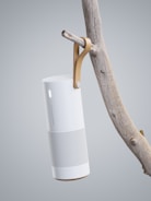 A sleek, modern cylindrical speaker hangs from a branch using a leather strap. The speaker is designed with a minimalistic style, combining white and grey colors with a perforated lower section for sound output. The branch has a natural, rustic appearance, contrasting with the speaker's contemporary design.