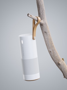 A sleek, modern cylindrical speaker hangs from a branch using a leather strap. The speaker is designed with a minimalistic style, combining white and grey colors with a perforated lower section for sound output. The branch has a natural, rustic appearance, contrasting with the speaker's contemporary design.