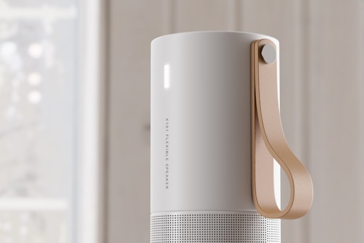 A sleek, cylindrical speaker with a matte white finish and a perforated lower section. It features a tan leather strap attached to the side for portability. The speaker has a minimalistic design with a small vertical light on the upper section.