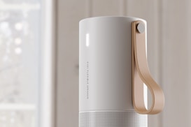 A sleek, cylindrical speaker with a matte white finish and a perforated lower section. It features a tan leather strap attached to the side for portability. The speaker has a minimalistic design with a small vertical light on the upper section.