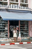 A storefront with a window display of various ceramic vases, featuring intricate blue patterns against a white background. Two people stand in front of the shop; an adult in a light-colored dress and hat, holding a tablet or book, and a child wearing a pink dress and blue hoodie. The shop has an awning above with a soft blue hue and there is a 30% sale sign in the window.