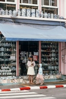 A storefront with a window display of various ceramic vases, featuring intricate blue patterns against a white background. Two people stand in front of the shop; an adult in a light-colored dress and hat, holding a tablet or book, and a child wearing a pink dress and blue hoodie. The shop has an awning above with a soft blue hue and there is a 30% sale sign in the window.