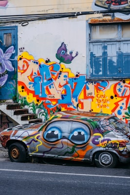 A vibrant and colorful graffiti scene features a car parked alongside a wall. The car is heavily painted with large, cartoonish eyes on the side, and swirling patterns in various colors. The wall behind it is covered in elaborate street art, including bold lettering and a purple creature with big eyes. The scene is urban and artistic, with an old staircase and blue window shutters adding to the background's texture.