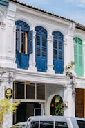 A traditional building facade with intricately decorated arched windows painted in a deep blue, contrasting with the white exterior. The shuttered windows are partly open, allowing a glimpse inside. A sign with Thai text hangs beside the entrance to a cafe named 'Chee Chee', indicated by a neon sign in the window. Overhead wires and a parked vehicle are visible in the foreground.