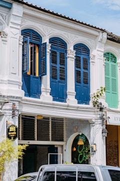 A traditional building facade with intricately decorated arched windows painted in a deep blue, contrasting with the white exterior. The shuttered windows are partly open, allowing a glimpse inside. A sign with Thai text hangs beside the entrance to a cafe named 'Chee Chee', indicated by a neon sign in the window. Overhead wires and a parked vehicle are visible in the foreground.