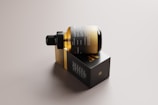 A sleek, amber glass bottle with a dropper cap is resting on its side on top of a rectangular black box. The packaging features minimalist design elements with gold and white text, conveying a sense of luxury and sophistication. The background is a neutral tone, highlighting the elegant design of the bottle and box.
