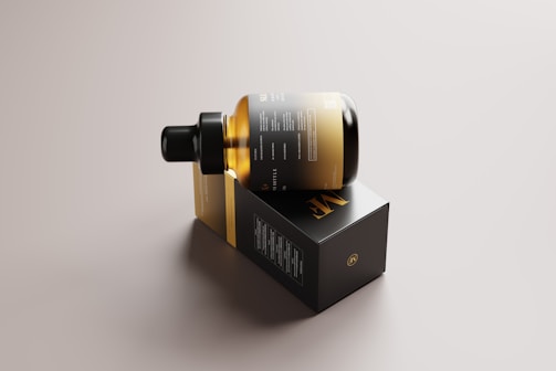 A sleek, amber glass bottle with a dropper cap is resting on its side on top of a rectangular black box. The packaging features minimalist design elements with gold and white text, conveying a sense of luxury and sophistication. The background is a neutral tone, highlighting the elegant design of the bottle and box.