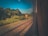 Railroad tracks extend alongside lush greenery, visible through a vehicle window. A sign marked with 'T' and 'Loop Cleared' stands by the tracks amidst dense foliage, highlighting a serene natural landscape under a clear blue sky.