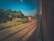 Railroad tracks extend alongside lush greenery, visible through a vehicle window. A sign marked with 'T' and 'Loop Cleared' stands by the tracks amidst dense foliage, highlighting a serene natural landscape under a clear blue sky.