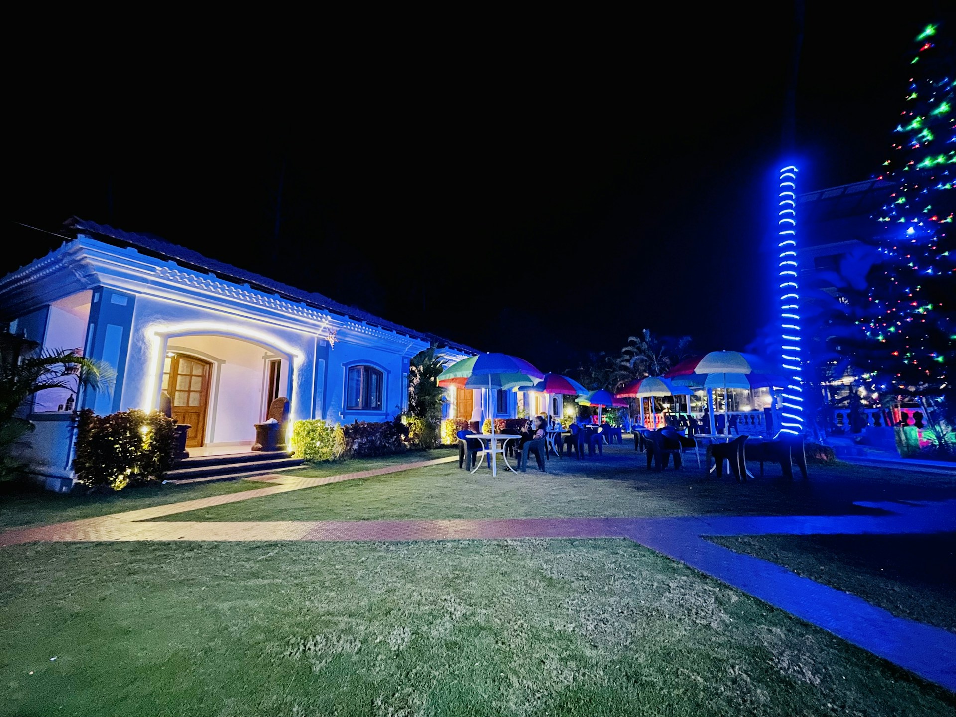 A well-lit house with decorative outdoor lighting stands in the foreground, with a garden featuring several tables and chairs under colorful umbrellas. A glowing, spiral light decorates a nearby tree or pole, adding to the festive ambiance. The scene suggests an evening event or gathering.