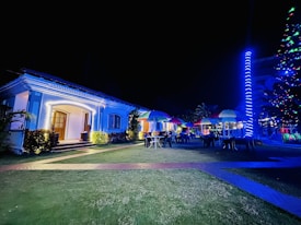A well-lit house with decorative outdoor lighting stands in the foreground, with a garden featuring several tables and chairs under colorful umbrellas. A glowing, spiral light decorates a nearby tree or pole, adding to the festive ambiance. The scene suggests an evening event or gathering.