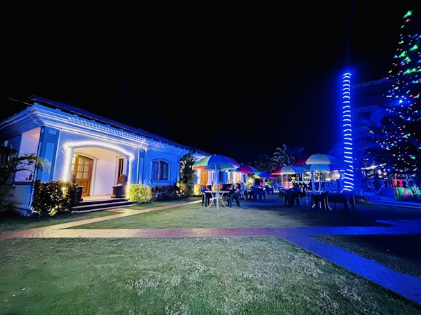 A well-lit house with decorative outdoor lighting stands in the foreground, with a garden featuring several tables and chairs under colorful umbrellas. A glowing, spiral light decorates a nearby tree or pole, adding to the festive ambiance. The scene suggests an evening event or gathering.