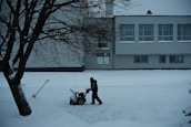 A solitary figure operates a snowblower on a snow-covered path in front of a large building. The scene is framed by a leafless tree on the left, with a shovel embedded in the snow nearby. The building has multiple windows with some snow accumulation on the sills, suggesting recent snowfall.