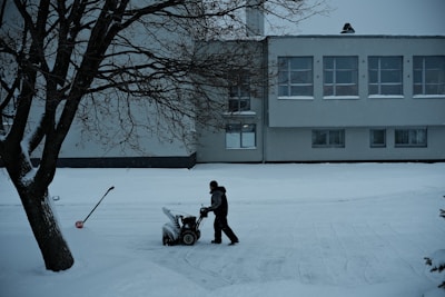 A solitary figure operates a snowblower on a snow-covered path in front of a large building. The scene is framed by a leafless tree on the left, with a shovel embedded in the snow nearby. The building has multiple windows with some snow accumulation on the sills, suggesting recent snowfall.