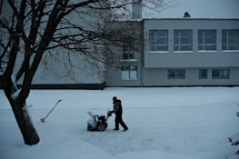 A solitary figure operates a snowblower on a snow-covered path in front of a large building. The scene is framed by a leafless tree on the left, with a shovel embedded in the snow nearby. The building has multiple windows with some snow accumulation on the sills, suggesting recent snowfall.