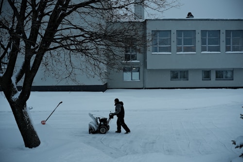 A solitary figure operates a snowblower on a snow-covered path in front of a large building. The scene is framed by a leafless tree on the left, with a shovel embedded in the snow nearby. The building has multiple windows with some snow accumulation on the sills, suggesting recent snowfall.