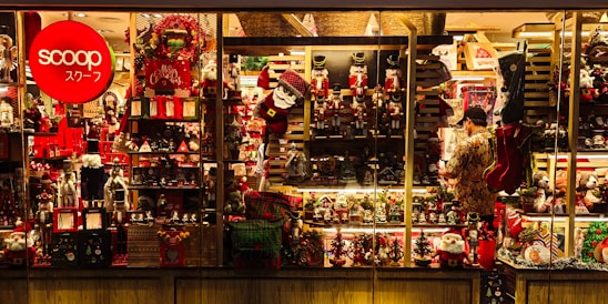 A brightly lit shop window displays a variety of Christmas-themed decorations, including nutcrackers, Santa figures, wreaths, and stockings. The display is richly colored with reds and greens, enhanced by warm lighting. A person stands inside, arranging items on the shelves.