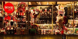 A brightly lit shop window displays a variety of Christmas-themed decorations, including nutcrackers, Santa figures, wreaths, and stockings. The display is richly colored with reds and greens, enhanced by warm lighting. A person stands inside, arranging items on the shelves.