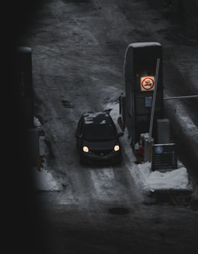 A car is passing through a snow-covered toll booth area at night. The road is icy and surrounded by snow, and there is a visible sign indicating no-entry for trucks.