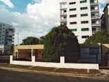 A residential street view featuring a tall white building with multiple balconies and green-tinted windows. In front of the building is a one-story beige house with a metal fence and a large tree in the front yard. The scene is flanked by other buildings, and the sky is partly cloudy with patches of blue.