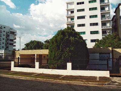 A residential street view featuring a tall white building with multiple balconies and green-tinted windows. In front of the building is a one-story beige house with a metal fence and a large tree in the front yard. The scene is flanked by other buildings, and the sky is partly cloudy with patches of blue.