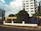 A residential street view featuring a tall white building with multiple balconies and green-tinted windows. In front of the building is a one-story beige house with a metal fence and a large tree in the front yard. The scene is flanked by other buildings, and the sky is partly cloudy with patches of blue.