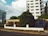 A residential street view featuring a tall white building with multiple balconies and green-tinted windows. In front of the building is a one-story beige house with a metal fence and a large tree in the front yard. The scene is flanked by other buildings, and the sky is partly cloudy with patches of blue.