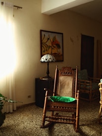 A room featuring a wooden rocking chair with a green cushion, positioned near a stained-glass table lamp and a piece of abstract wall art. Sunlight filters through white curtains, casting a warm glow over the speckled tile floor. A second seating area with a bamboo chair is partially visible.